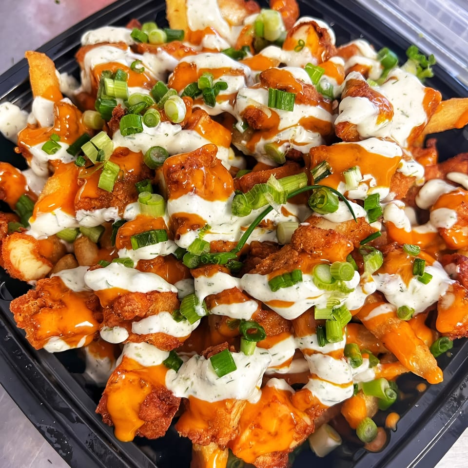 Buffalo chicken fries topped with ranch dressing and green onions in a black takeout container.