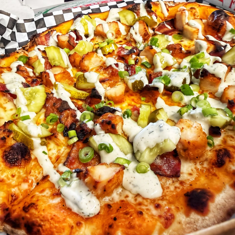 Close-up of a pizza topped with chicken, bacon, pickles, ranch, and green onions in a box.